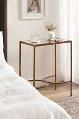 Bronze metal side table with a rectangular top, holding a glass of water, white ceramic vase with dried flowers, and eyeglasses, positioned next to a bed and sheer curtain.