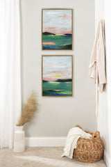 Two framed abstract landscape paintings with green and blue foregrounds and pinkish skies, hung vertically on a light wall beside beige curtains, a woven basket, and a vase with pampas grass.