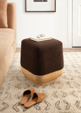 Square ottoman with dark brown textured fabric top and light wood base, placed on a patterned beige rug beside a tan sofa, with glasses and a book on top.