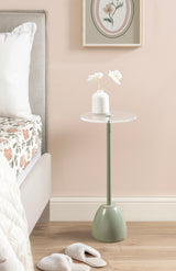 Minimalist side table with a round, clear top and olive green angled base next to a bed with floral bedding and white slippers on wood floor.