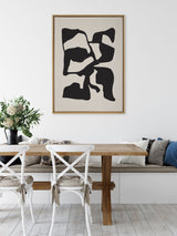 Abstract black irregular shapes on a tan canvas, framed in light wood and hung above a wooden dining table with white chairs and a floral centerpiece.