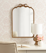 Arched wall mirror with a ribbon-shaped topper and wooden frame above a desk holding a bronze table lamp and a vase with pink flowers.
