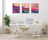 Three framed abstract canvases with vibrant brush strokes in pink, blue, orange, and white, arranged horizontally above a minimalist white desk with a beige chair and plants on each side.