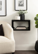 Black rectangular floating side table with rounded edges mounted on a white wall, holding a black vase with greenery and a white textured jar, above stacked books below.