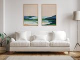 Neutral-toned living room with a white cushioned sofa, two abstract ocean-themed canvas prints framed in light wood, a potted plant, and a tall floor lamp with a beige shade.