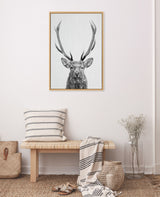 Black and white framed canvas of a red deer head with large antlers hanging above a simple wooden bench with striped pillow and draped blanket in a minimalist room.