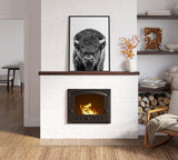 Black and white framed portrait of a bison centered above a lit fireplace in a white brick wall, flanked by a vase with orange flowers and neatly stacked firewood.