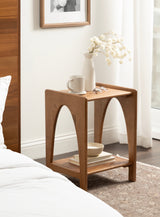 Light brown wooden side table with rounded arches on each side, featuring a white vase with flowers and a mug on top, next to a bed and window with curtains.