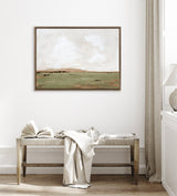 Framed canvas artwork depicting a soft, muted landscape with light beige sky and greenish-brown terrain, hanging above a wooden bench draped with a light fabric and an open book.