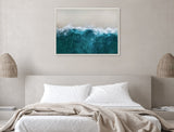 Framed aerial view of ocean waves crashing onto a sandy beach, featuring deep blue water and white foam, hanging above a neutral-toned bed with beige linens.