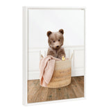 Framed canvas artwork of a brown bear cub sitting inside a woven laundry basket with a pink knitted blanket and a small yellow rubber duck, against a white paneled wall and wooden floor.
