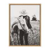 Black and white photo of a woman wearing a hat standing beside a white horse in a field, holding a small flower in her outstretched hand.