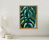 Framed painting of large green monstera leaves with dark and light green shades against a pale blue background on a white wall above a table with a white textured lamp and a small round white candle.