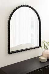 Arched black wall mirror with beaded frame hung above a black cabinet, next to a candle and a vase with green foliage on a white wall.