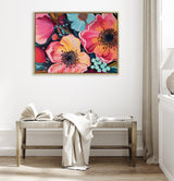 Framed canvas artwork of large stylized flowers in vibrant pink, peach, and blue tones with detailed centers, hanging above a wooden bench with neutral textiles in a bright room.