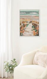 Framed canvas painting of a sandy boardwalk leading through orange flowers toward a teal and pink cloudy beach horizon, hung on a white wall above a cream armchair.