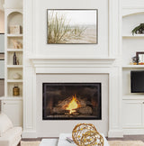 Neutral-toned living room with white fireplace mantel and burning fire, topped by framed print of sandy beach grass under a pale sky.