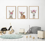 Three framed animal portraits with flower crowns: a rabbit, a fawn, and an alpaca, hung above children's toys and a chevron cushion on a white wall.