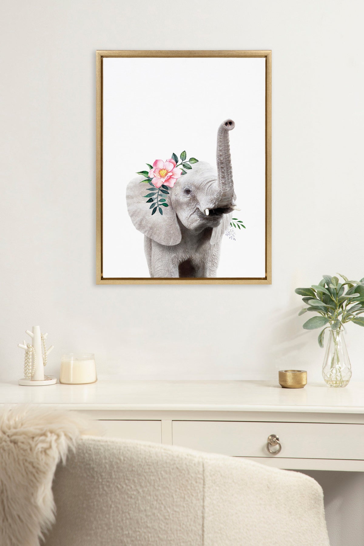 Framed canvas print of a gray baby elephant raising its trunk adorned with a pink flower crown and green leaves on a white wall above a white drawer with plants and decor.