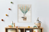 Framed canvas of a pastel teal and peach striped hot air balloon carrying an elephant and lion in a wicker basket, hung above a wooden shelf with toys and colorful pom-pom decor on a white wall.