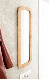 Tall, narrow rectangular mirror with rounded corners and a woven light wood frame mounted on patterned beige wallpaper.