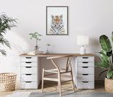 Framed close-up portrait of a calm tiger with white and orange fur, centered above a modern light wood desk with white drawer units, natural wood chair, and green plants.