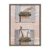Two giraffes peeking out from flower-filled window boxes with gray shutters on a pale pink stucco wall, framed in simple gray framing.