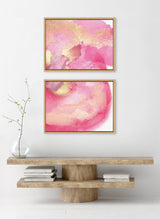 Two framed abstract canvases with flowing pink, gold, and white patterns hang vertically above a natural wood console table with a round base, decorated with a glass vase and stacked books.
