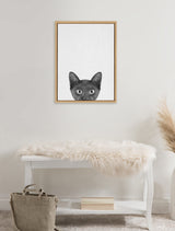 Black kitten's head peeking up from bottom of a beige canvas within a natural wood frame, above a white bench draped with a fluffy cream fur throw.