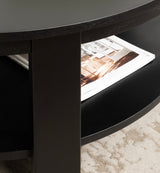Close-up of a black round wood coffee table with a thick central leg and a lower shelf holding a magazine, set on a beige textured rug.