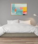 Framed abstract canvas by Amy Lighthall with soft yellow, teal, and coral blocks, hanging above a white-bed with gray wall and floor lamp on the side.