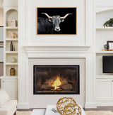 Framed canvas of a black longhorn bull with white facial markings and curved horns on a black background, hanging above a white fireplace with a burning fire.