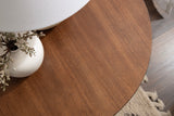 Close-up of a round wood side table top with a warm brown finish, featuring a white vase with dried flowers and a lamp base partially visible.