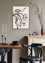 Black and white floral line art print in a beaded wooden frame hanging above a beige mantel with pottery and branches, beside a wooden dining table and black chairs.