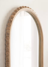 Arched wall mirror with a beige frame featuring a row of rounded bead-like accents along the outer edge, reflecting white curtains in soft natural light.