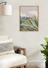 Abstract mountain landscape in muted green, blue, and beige tones with textured clouds, framed and hung on a light gray wall above a beige couch and near a green plant.