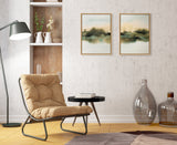 Two framed abstract landscape paintings with soft green and beige tones hang above a tan cushioned lounge chair, round black side table, and two large glass bottles on a wooden floor.