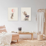 Two framed canvas prints of rabbits on a white wall, one holding a pink balloon, the other wearing star-shaped sunglasses, above a light wood bench with lavender and candles.