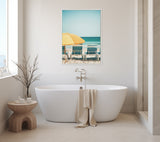White oval freestanding bathtub with beige towel draped over edge, beside a round wooden stool and a framed canvas of yellow beach umbrella and striped chairs on a light wall.