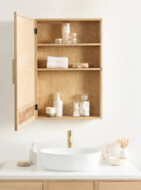 Open wooden vanity cabinet with two shelves holding toiletries and towels above a white oval sink and gold faucet on a white countertop against a plain white wall.