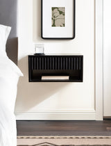 Black floating wood side table with vertical slats on front panel and open lower shelf, mounted on white wall next to bed with grey headboard.
