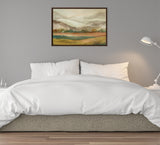 Abstract mountain landscape painting in muted greens, browns, and golds, framed and hung on a grey wall above a white bed with a lamp on a nightstand to the right.