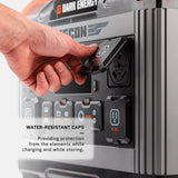 Close-up of a hand lifting a water-resistant protective cap on the Recon 2000 power station's AC outlet, showing black textured cover and multiple ports.
