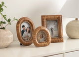 Three woven rattan picture frames in oval, small oval, and rectangular shapes arranged on a white dresser with a ceramic vase and wooden lamp nearby.
