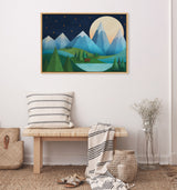 Framed canvas painting depicting blue and green geometric mountains under a large pale moon with dark sky and golden stars, hanging above a wooden bench with striped pillows and throw.