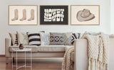 Neutral-toned living room with beige sofa, patterned black and white pillows, chunky knit throw, and three framed minimalist prints: three cowboy boots, stylized 'HOWDY', and a cowboy hat.