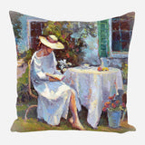 Painting on pillow of a woman in a white dress and wide-brimmed hat sitting barefoot reading beside a round table with a white cloth, near a green shutter and flowers.
