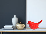 Bright red ceramic bird figurine with smooth, curved surface displayed on white marble shelf alongside a gold-colored rounded jar, minimalist gray vase, and stacked books.