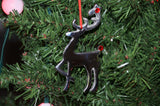 Black reindeer ornament with red gem eyes and nose hanging by red string on green artificial Christmas tree branches with pink lights in background.