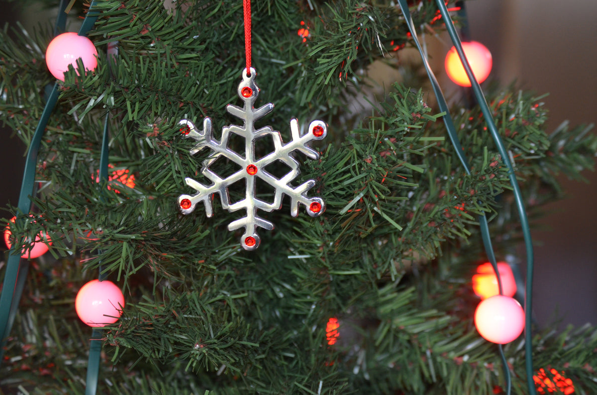 Silver snowflake ornament with red gems hanging on green Christmas tree branches with pink round string lights.